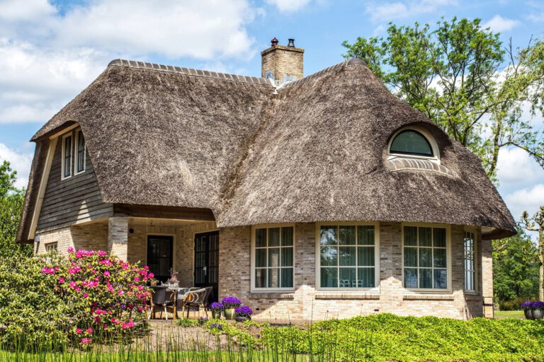 Traditional house with rustic thatched roof in a sunny day. Trees, flowers and bright blue sky with clouds on background. Giethoorn, Netherlands